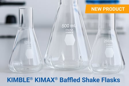 Three KIMBLE® KIMAX® Baffled Shake Flasks (125 mL, 250 mL, and 500 mL) made of clear borosilicate glass are displayed on a laboratory bench with a blurred microscope and lab equipment in the background. An orange label in the top right corner reads “NEW PRODUCT,” and bold blue text at the bottom says “KIMBLE® KIMAX® Baffled Shake Flasks.”